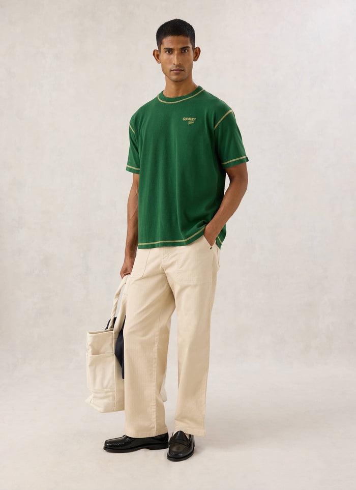 Model wearing the forest green Guinness x Percival T-shirt, paired with light trousers and holding a tote bag.